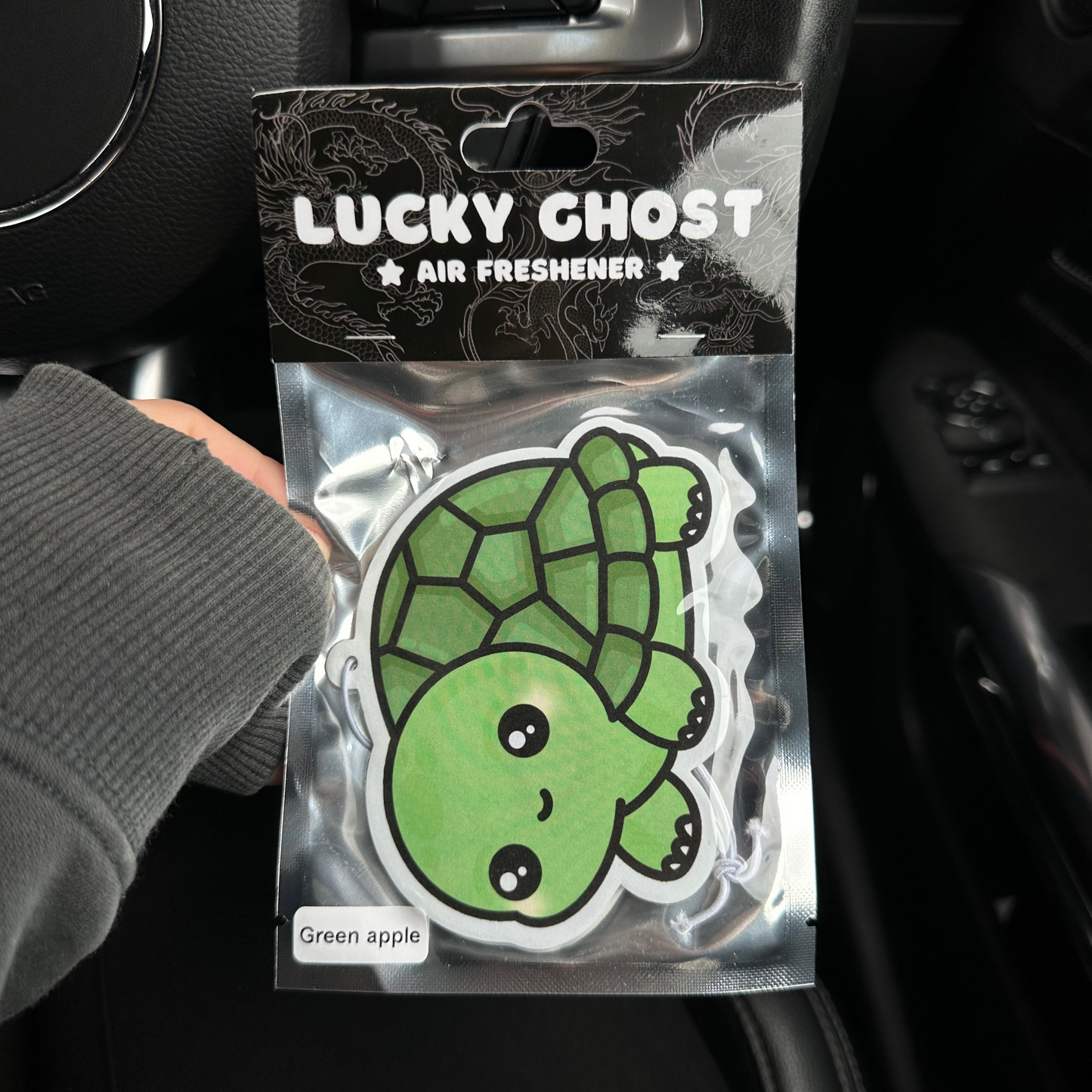 Lucky Ghost air freshener packaging with a green turtle design held by a hand inside a car.