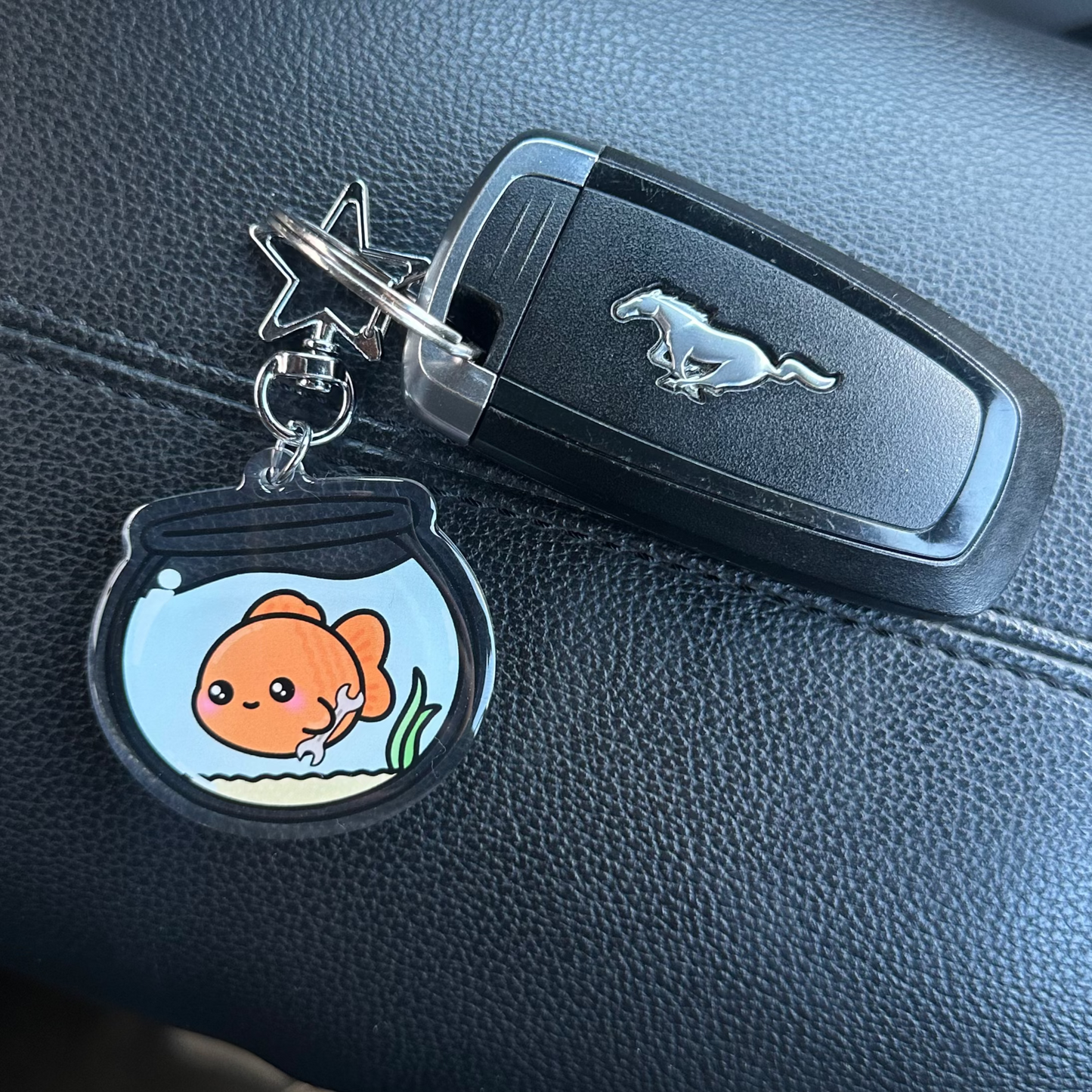Keychain with a cartoon goldfish in a fishbowl attached to a car key on a black leather surface.