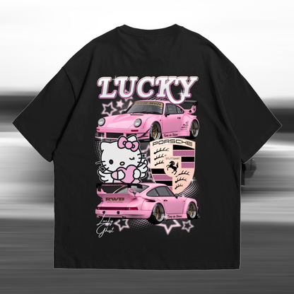A black short-sleeve graphic T-shirt shown flat against a blurred gray studio background. The shirt features a large front print with the word 'LUCKY' in pink outlined letters above two pink cars, a Hello Kitty character, and a Porsche crest motif.