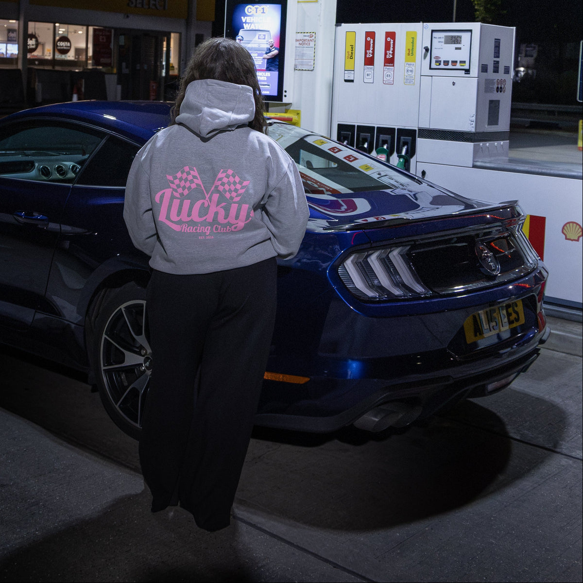 A person wearing a light gray hoodie with pink text and black pants stands at a gas station pump next to a dark blue sports car under bright overhead lights at night.