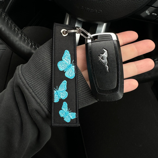 Keychain with butterfly design held by a hand in front of a car steering wheel.
