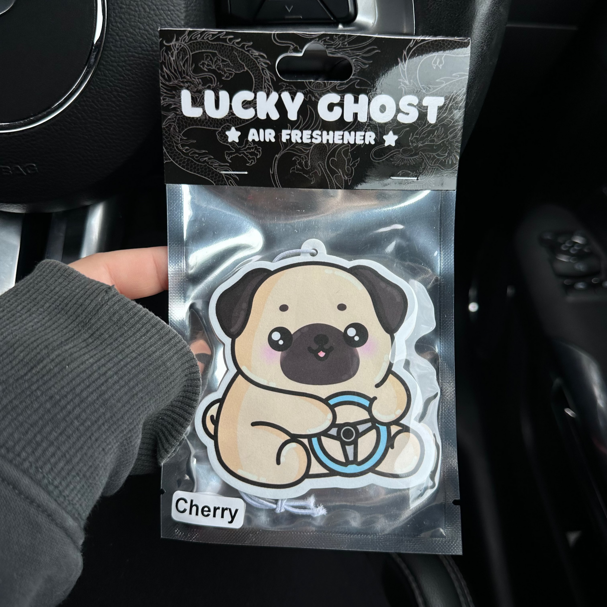 Lucky Ghost air freshener packaging with a pug design held in front of a car's dashboard.