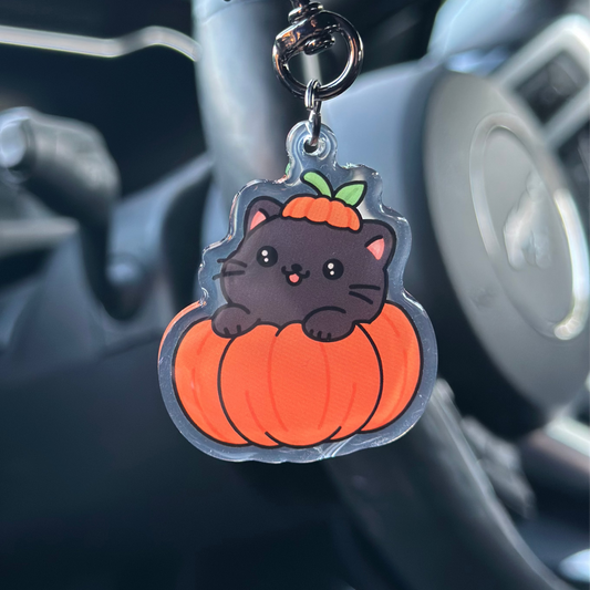 Keychain with a black cat on an orange pumpkin hanging from a car's keychain holder.