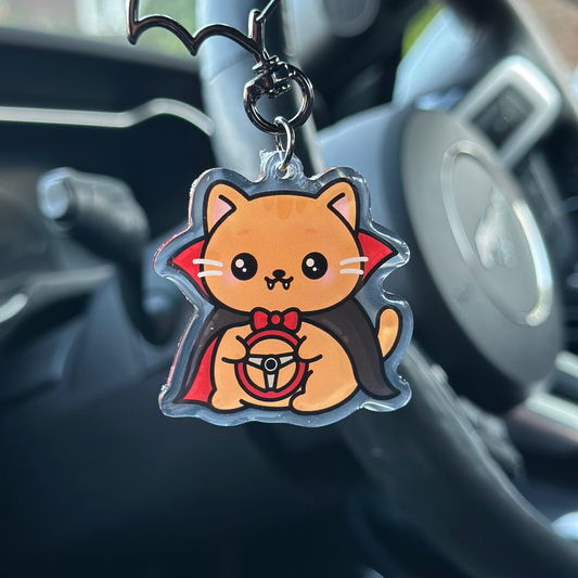 Keychain shaped like a cartoon cat in a vampire costume hanging from a car's keychain holder.