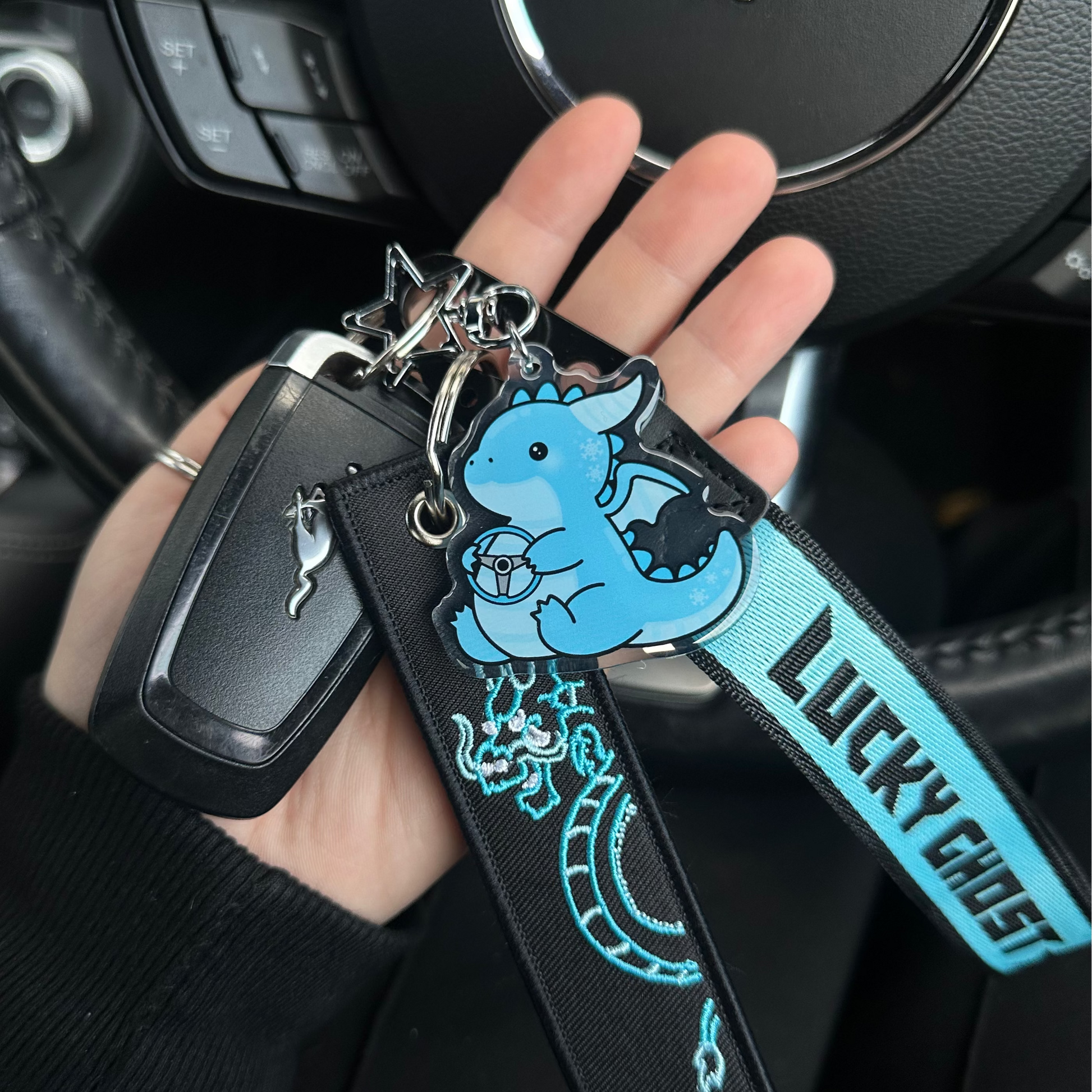 Hand holding a keychain with a blue ghost design in front of a car steering wheel with a Mustang logo.