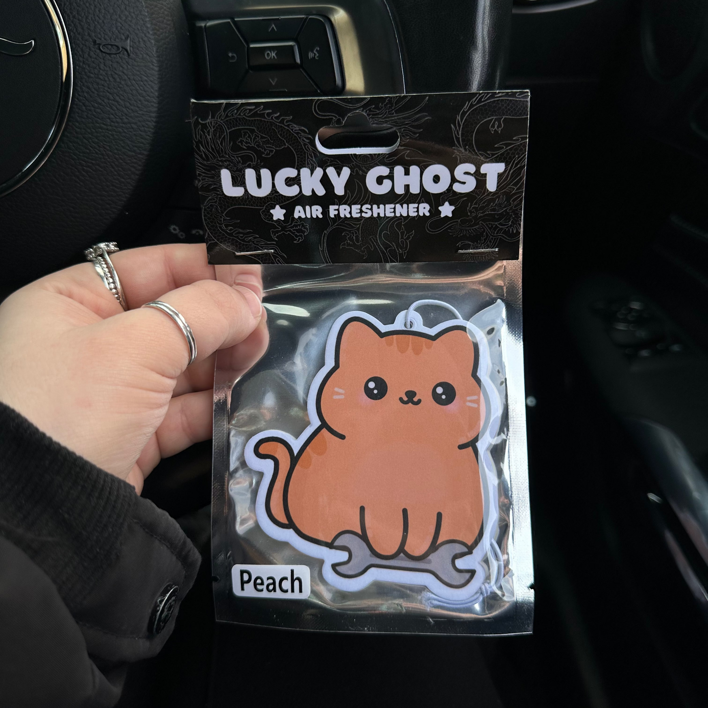 Hand holding a 'Lucky Ghost' air freshener with a peach cat design inside a car.