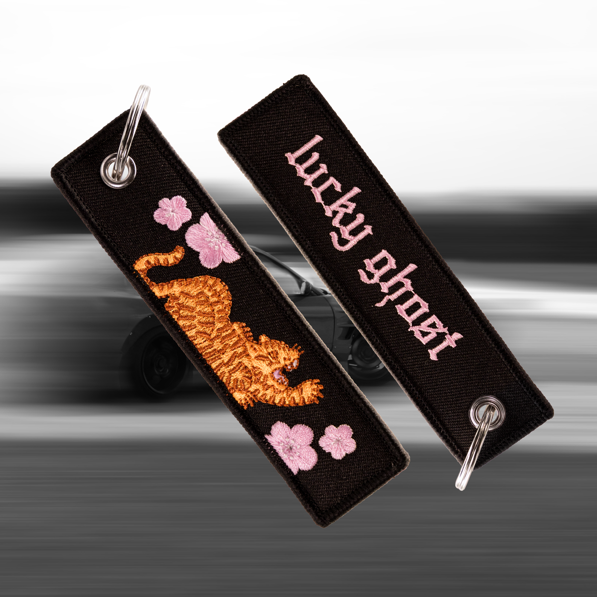 Two black embroidered keychain tags with metal split rings, shown against a blurred gray background. One tag features a stylized orange tiger and pink cherry blossoms, while the other features the text 'lucky ghost' in pink gothic lettering.